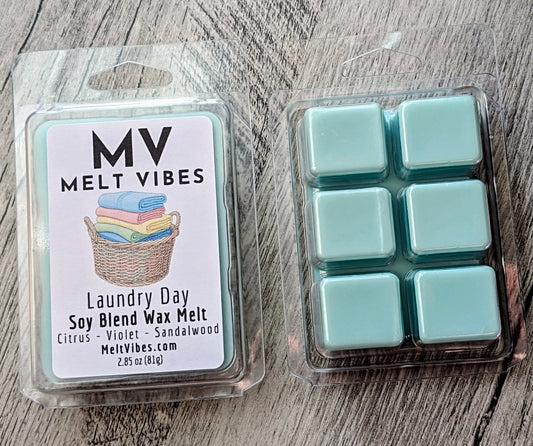 Melt Vibes Laundry Day soy blend wax melt in a clear clamshell package with a white label, shown next to six mint green wax melts in a clear plastic tray on a gray wood-textured background.