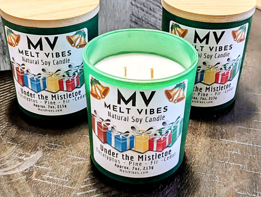 Three Melt Vibes 'Under the Mistletoe' natural soy candles in green glass jars with wooden lids, with one candle in the center open to show the white wax and wicks, set on a dark wooden surface.