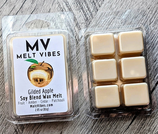 Two packages of MV Melt Vibes Gilded Apple soy blend wax melt shown on a gray wood background. One package is the retail clamshell with a white label, and the other shows six individual cream-colored wax melts inside clear plastic packaging.