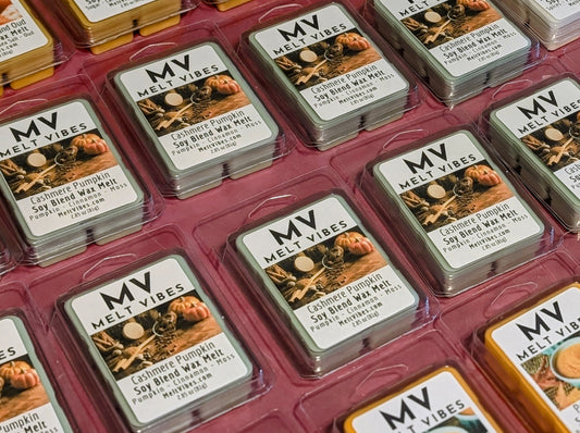 Multiple small rectangular wax melts in clear plastic clamshell tins arranged in a pink tray. The front tins show a white label with the brand name MV Melt Vibes and a pumpkin-themed scent label, while other tins in the background show different label colors.