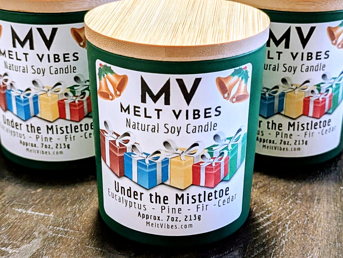 Three green natural soy candles with bamboo wood lids are arranged on a dark wooden surface. The front candle is in sharp focus, showing a white label with the brand 'Melt Vibes' and the scent 'Under the Mistletoe', decorated with illustrations of Christmas gifts.