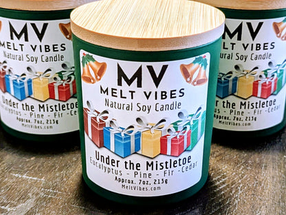 Three green natural soy candles with bamboo wood lids are arranged on a dark wooden surface. The front candle is in sharp focus, showing a white label with the brand 'Melt Vibes' and the scent 'Under the Mistletoe', decorated with illustrations of Christmas gifts.